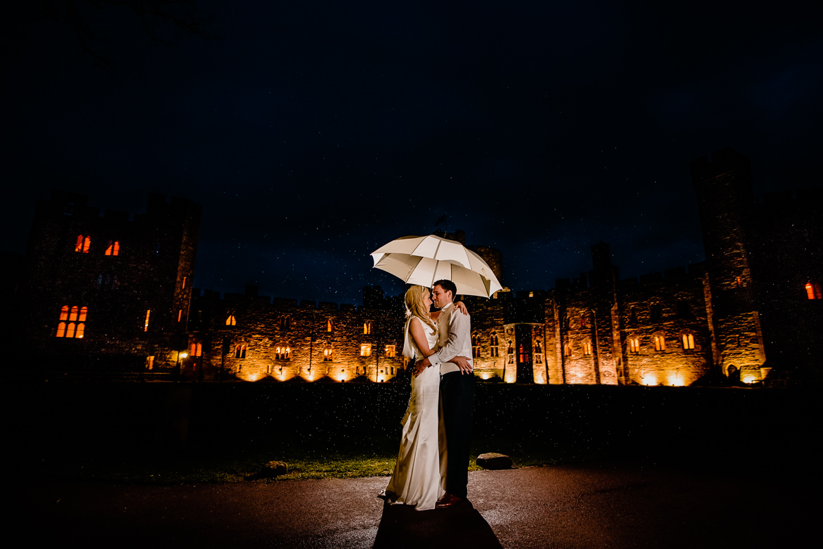 Backlit rain with the bride and groom holding an umbrella in form of Peckforton Castle