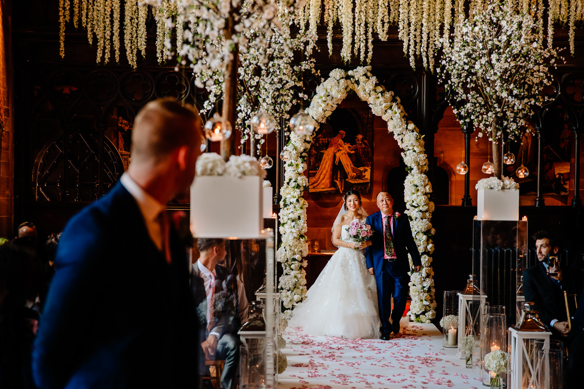 Bride walking down the wedding aisle with her father looked on by her husband to be in the great Hall at Peckforton Castle