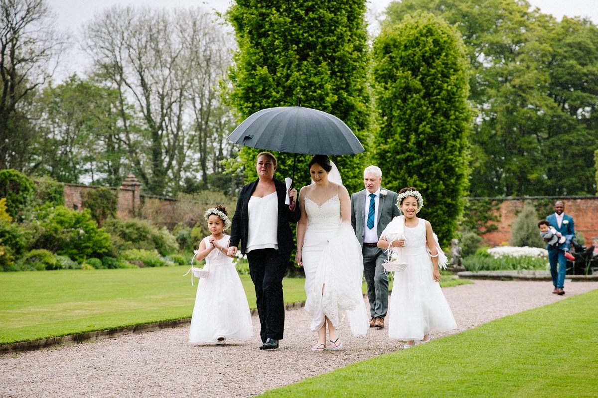 Wedding coordinator sheltering the bride as she makes her way to the alcove for the outdoor wedding ceremony at Arley Hall