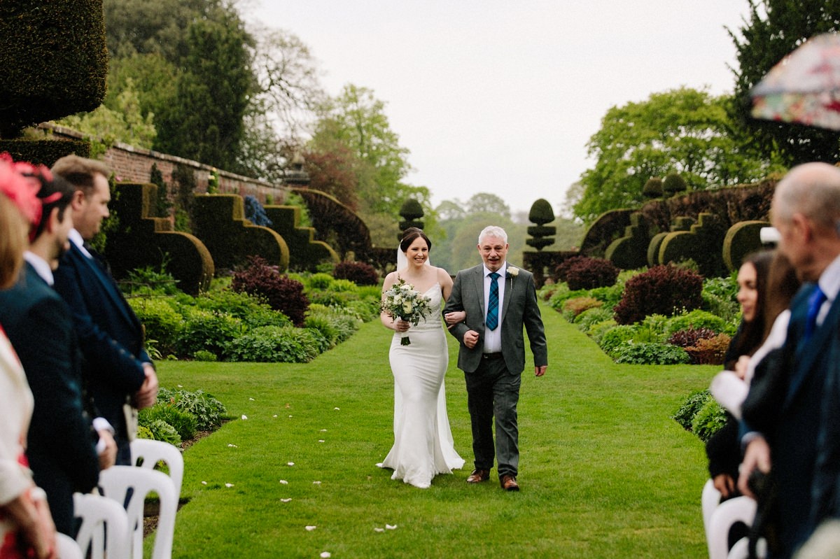 Bride walking down the aisle with her father for the outdoor wedding ceremony at Arley Hall