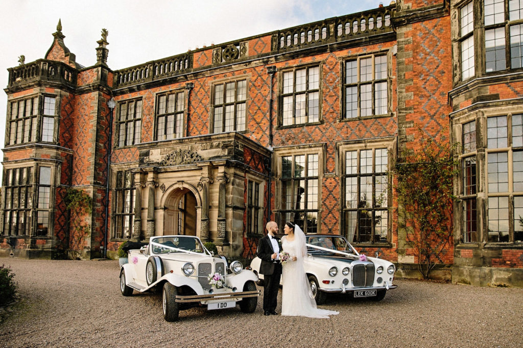 Bride and groom in front of the stately Arley Hall with two stunning wedding cars